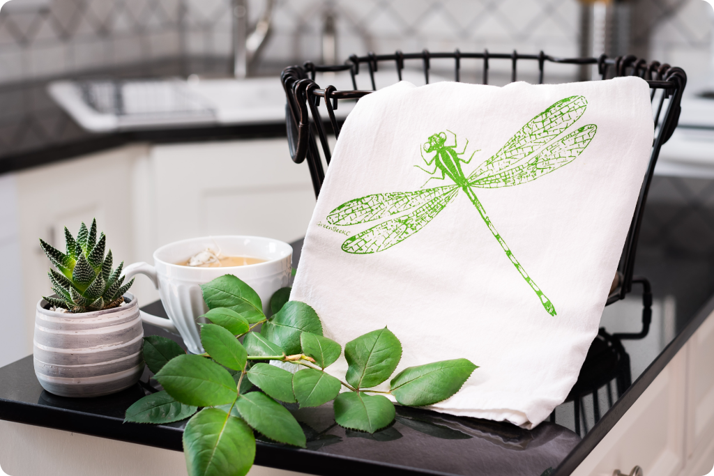 White towel with green dragonfly design on a black tray with plants and a cup.