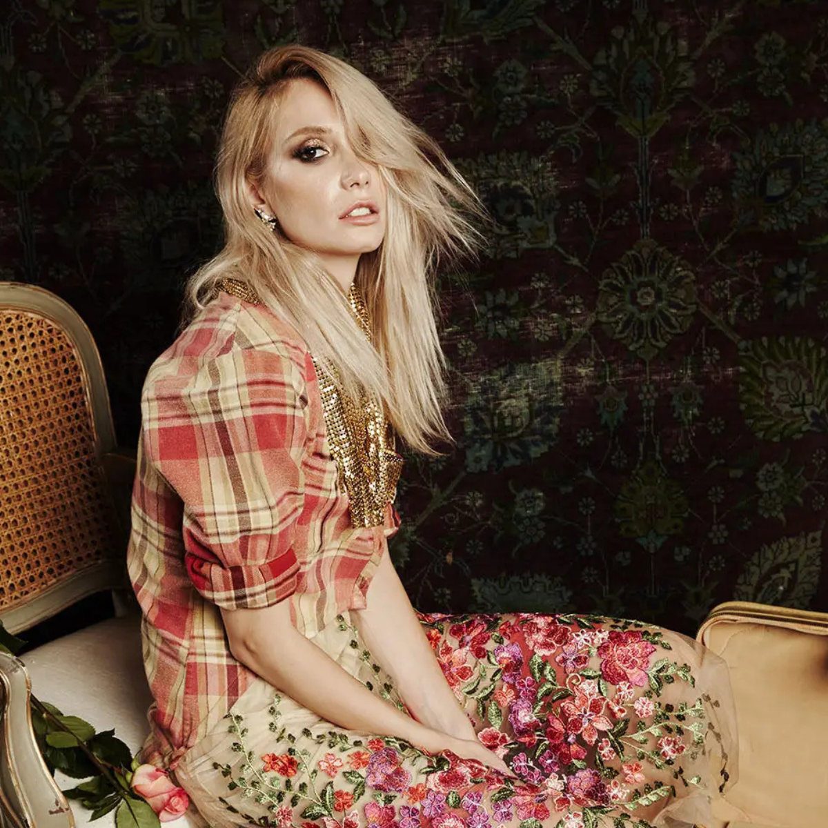 Woman sitting on a decorative chair wearing a plaid shirt and floral dress against a dark, patterned wall.