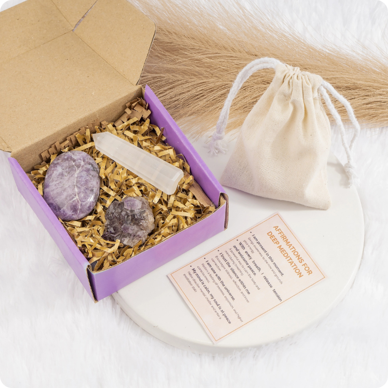 A crystal gift set in a purple box with shredded paper, including two polished purple stones, a white crystal pointer, and a small beige drawstring pouch, arranged on a white surface with a card beside it.