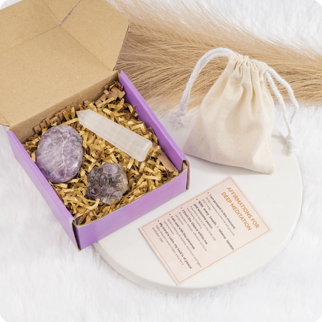 A crystal gift set in a purple box with shredded paper, including two polished purple stones, a white crystal pointer, and a small beige drawstring pouch, arranged on a white surface with a card beside it.