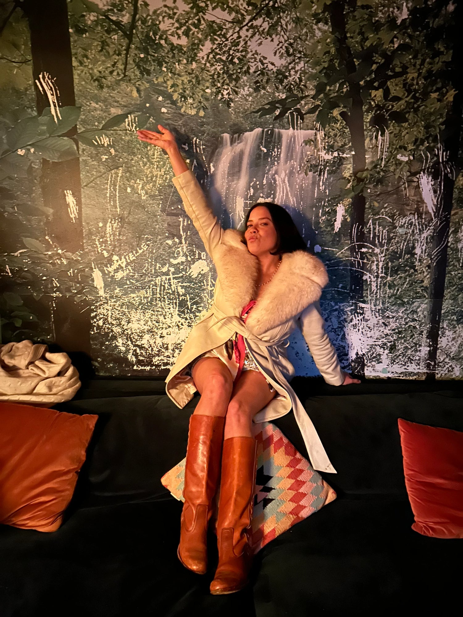 Umbelina in a fur coat and brown boots sitting on a couch with a scenic wall painting in the background