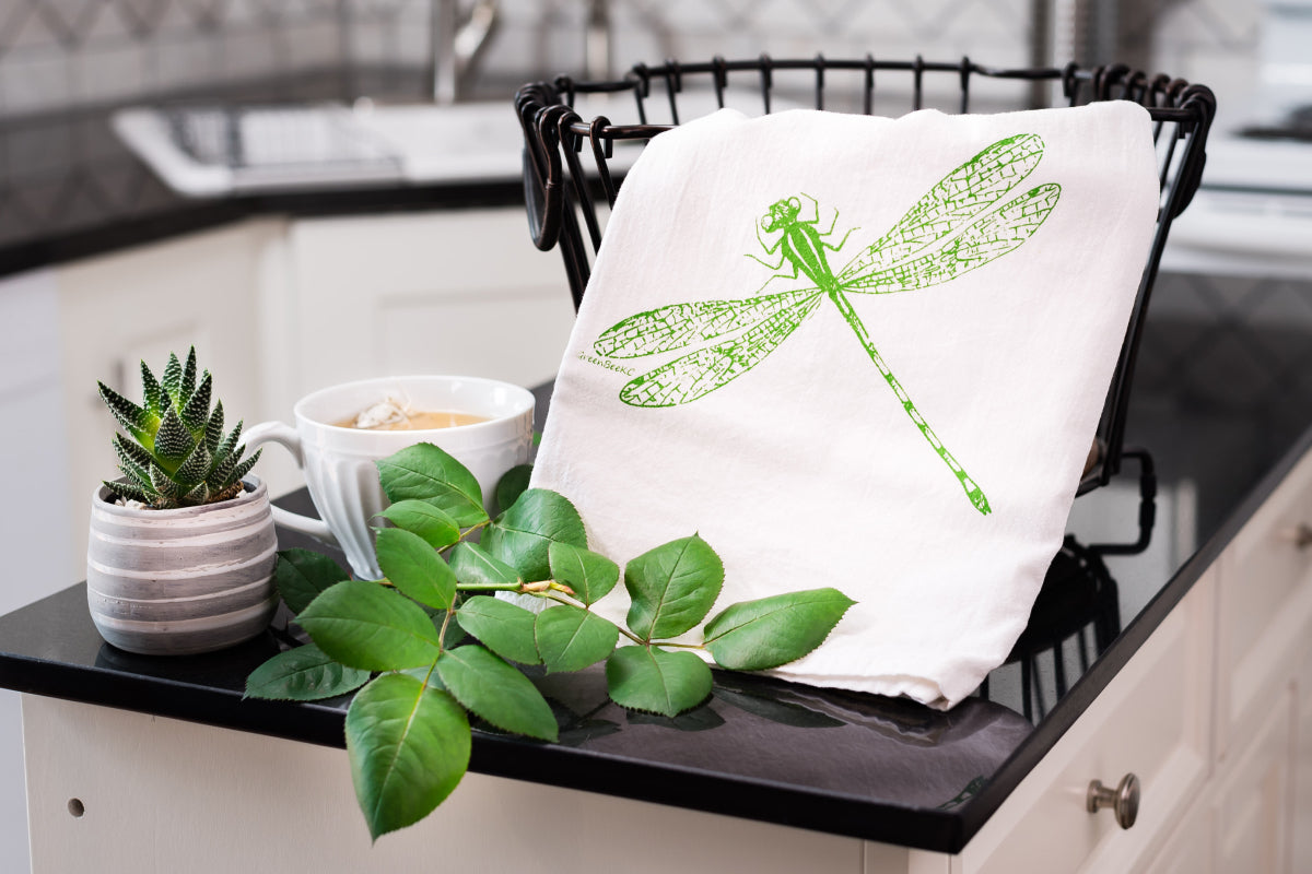 White towel with green dragonfly design on a kitchen counter with plants and a cup.
